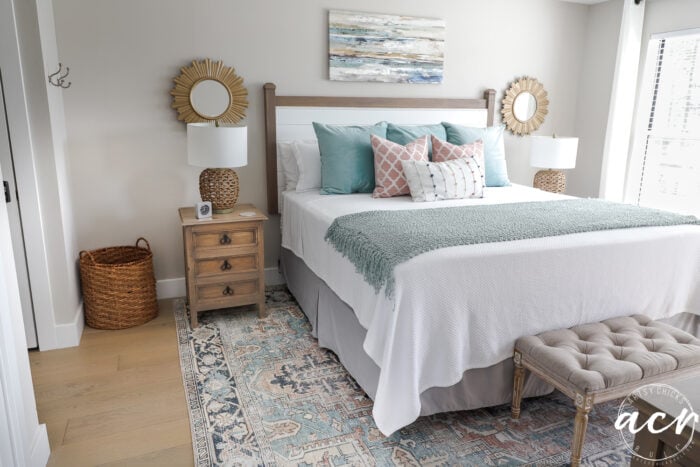 Kamar tidur dengan penutup putih dan bantal berwarna -warni, cetakan berwarna -warni di atas tempat tidur, cermin starburst emas di setiap sisi, karpet berwarna -warni
